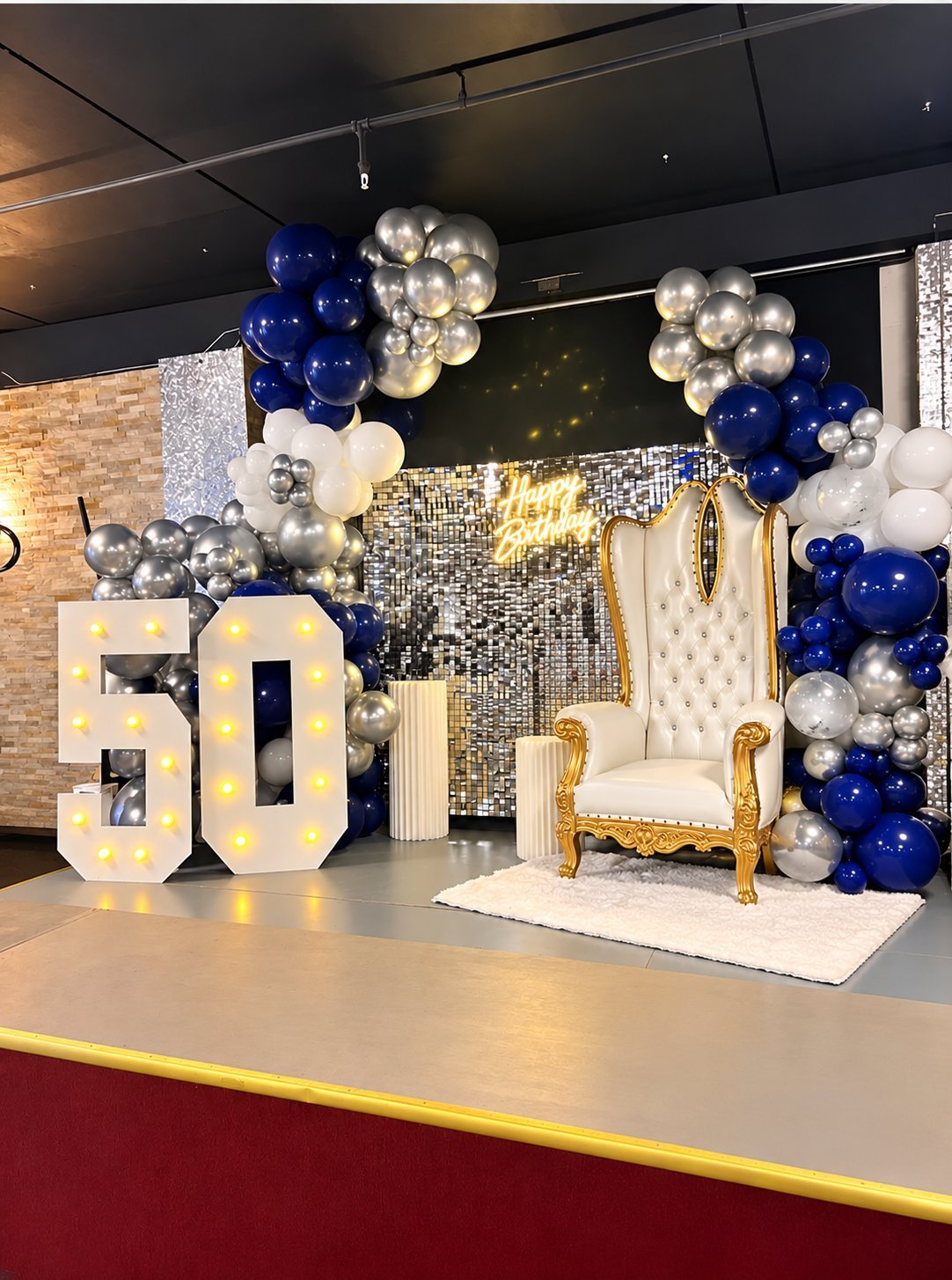 50th birthday party setup with illuminated number, white upholstered chair, silver and blue balloon garland, and sequin backdrop